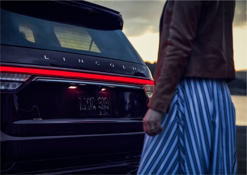A person is shown near the rear of a 2023 Lincoln Aviator® SUV as the Lincoln Embrace illuminates the rear lights | Eby Lincoln in Goshen IN